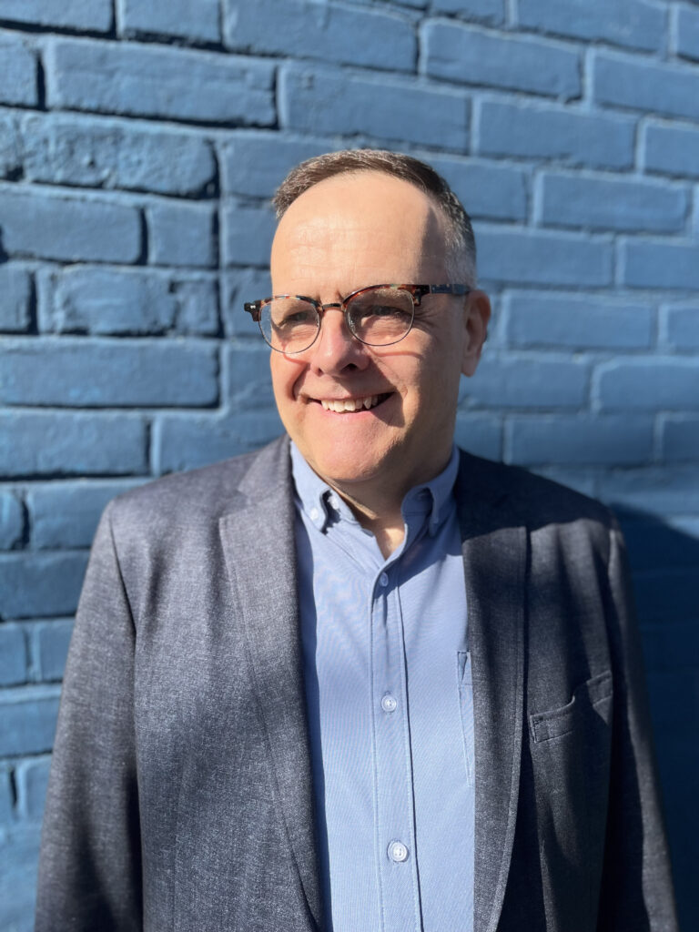 Photo of Matt smiling and looking to the side of the camera. He is wearing a light blue button up shirt, a grey blazer jacket and glasses. There is a brick wall behind him.